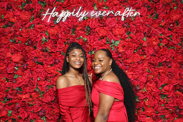 Two young women each in a red dress smiling in front of a red rose backdrop.